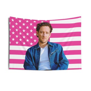 May include: A tapestry featuring a man in a denim jacket and white shirt, set against a pink and white American flag backdrop. The flag design includes white stars on a pink field and pink and white stripes.