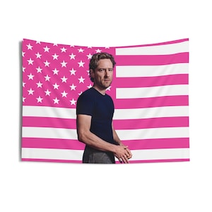 Puede incluir: Un tapiz de la bandera estadounidense rosa y blanca con un hombre con una camiseta azul marino. La bandera presenta estrellas blancas sobre un fondo rosa y rayas rosas y blancas. El hombre está de pie frente a la bandera.