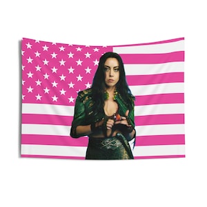 May include: A tapestry featuring a pink and white American flag design with a woman in the center. The woman is wearing a green jacket and a dark skirt. The tapestry is rectangular and hangs against a white background.