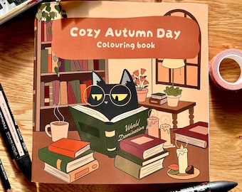 May include: A square colouring book titled Cozy Autumn Day Colouring book featuring an illustration of a black cat wearing glasses and reading a book labelled World Domination, surrounded by stacks of books, candles, and a mug.