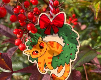Könnte beinhalten: Weihnachtsbaumschmuck aus Holz mit einer orangefarbenen Tabby-Katze mit roten Augen, in einem grünen Kranz mit roter Schleife. Der Schmuck hängt an einer rot-weiß gestreiften Schnur. Der Hintergrund zeigt rote Beeren und grüne und burgunderfarbene Blätter.