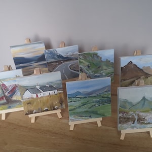May include: A collection of 10 small, framed paintings on easels. The paintings are all landscapes, featuring mountains, valleys, and seascapes. The paintings are all done in a realistic style, with a focus on detail and colour.
