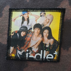 May include: A square patch with a photo of five people, with the word "idle" at the bottom. The patch has a black border with yellow and white colors in the photo. The people are wearing various outfits, including hats and shirts.