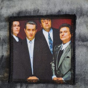 May include: A rectangular patch featuring a group of four men in suits. The image has a red background and a black border. The men are wearing suits and ties, with varying expressions. The patch is likely for clothing or accessories.