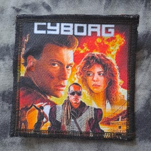 May include: Square patch with the word "CYBORG" in white block letters. The patch features a movie poster with three people in front of a fiery background. The patch has a black border and is on a gray and white patterned fabric.