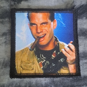 Op de afbeelding: Vierkante patch met zwarte rand, met een lachende man met een sigaar. De achtergrond is blauw. De man draagt een beige jas en een zwart shirt met een wit en rood ontwerp.