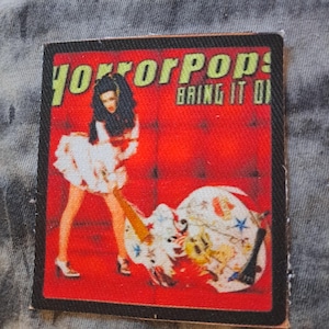 May include: A square patch with a red background featuring the text "horrorpops!" in yellow and "BRING IT ON" in white. The patch depicts a woman in a white dress and a broken guitar.