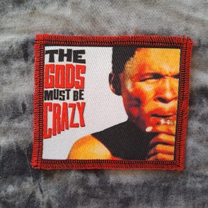 May include: Square patch with a red border and the text "THE GODS MUST BE CRAZY" in red block letters. The patch features a portrait of a man with an orange complexion.