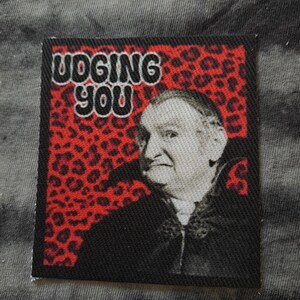 May include: A rectangular patch with the words "JUDGING YOU" in white letters. The background is red with black leopard spots. A black and white image of a person is in the center of the patch.