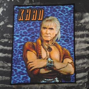 May include: Rectangular patch featuring a portrait of a man with silver hair and a stern expression. The patch has the word "KHAN" in orange letters at the top. The background is blue with a leopard print pattern. The man is wearing a brown and gold outfit.
