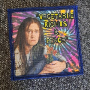Pode incluir: Um emblema de tecido quadrado com uma borda azul costurada, apresentando uma pessoa com cabelos longos e castanhos segurando um pequeno buquê de flores. O fundo é um padrão tie-dye colorido com o texto amarelo "VEGETABLE RIGHTS & PEACE".