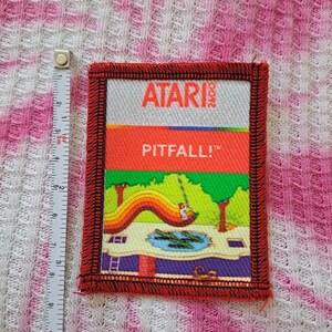 Może przedstawiać: Prostokątna naszywka Atari Pitfall! z czerwoną obw&oacute;dką. Naszywka zawiera logo Atari i tytuł gry pogrubioną czcionką. Kolorowa grafika przedstawia tęczę, drzewa i staw.