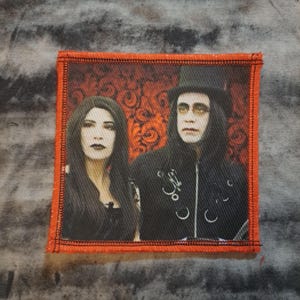 May include: A square patch with an orange border featuring a portrait of two people in gothic attire. The background is a dark red floral pattern. The subjects are dressed in black, with pale makeup and dark hair.