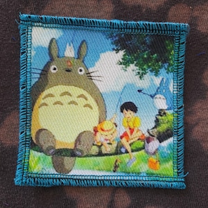 May include: Square patch featuring a scene from the animated film, My Neighbor Totoro. The patch depicts Totoro, a large gray forest spirit, with two children and other forest spirits in a colorful outdoor setting. The patch has a blue border.