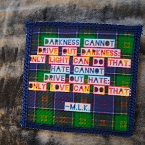 May include: Square patch with a blue border and a plaid pattern in green, purple, and blue. The patch features the quote "DARKNESS CANNOT DRIVE OUT DARKNESS: ONLY LIGHT CAN DO THAT. HATE CANNOT DRIVE OUT HATE: ONLY LOVE CAN DO THAT." -M.L.K.