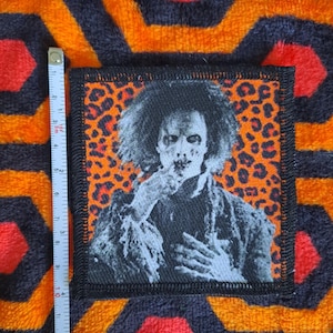 May include: A square patch featuring a black and white image of a character with wild hair and a distressed appearance. The patch has a black border and an orange leopard print background. The patch measures approximately 3 inches by 3 inches.
