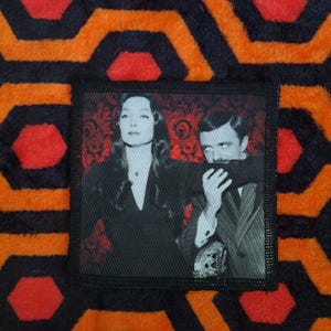 May include: A square patch featuring a black and white image of a man and woman. The patch is set against a patterned orange, black, and red background. The background has a repeating geometric design.
