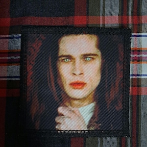 May include: Square patch featuring a portrait of a person with long, dark hair, pale skin, and striking blue eyes. The person has red lips and is set against a dark background. The patch is bordered with black stitching and is likely an iron-on or sew-on patch.
