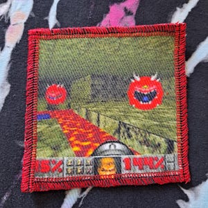 May include: Square patch with a red stitched border featuring a pixelated scene with a red lava path, green landscape, and two red demon-like creatures. The patch has a retro video game aesthetic.
