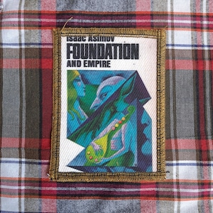 以下が含まれることがあります： アイザック・アシモフの「Foundation and Empire」の表紙アートをあしらった長方形のパッチ。パッチは白い背景に、青、緑、黄色の人物のカラフルなイラストが描かれています。タイトルは黒い文字です。