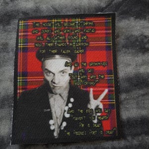 May include: A rectangular patch with a portrait of a man in a black jacket and a beret, set against a red and green plaid background. The patch includes white text with song lyrics and the phrase "The People's Poet is dead."