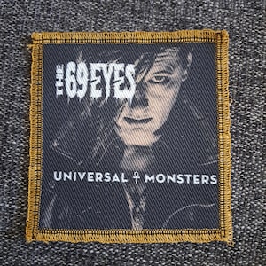 Könnte beinhalten: Ein quadratischer Stoffaufnäher mit goldfarbenem Stickrand, der ein schwarz-weißes Porträt einer Person mit dunklem Haar zeigt. Der Text THE 69 EYES ist oben in Weiß gedruckt, und UNIVERSAL MONSTERS ist unten in Weiß gedruckt.
