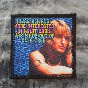 May include: Square patch with a portrait of a person with blonde hair and the text "I WAS ALWAYS MORE INTERESTED IN WHAT BARK WAS MADE OUT OF ON A TREE" in colorful letters. The background has a blue leopard print.