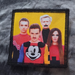 Peut inclure: Un écusson carré avec un fond jaune représentant quatre personnes. La personne au centre porte un pull rouge avec un motif Mickey Mouse. L'écusson est bordé d'une bordure noire.