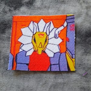 May include: A square fabric patch featuring a stylized robot design. The robot has a yellow head, white flower-like details, and a red chest. The background is orange and purple, with a gray fabric background.