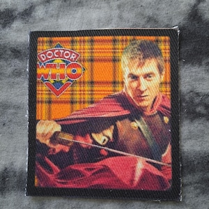 May include: A rectangular patch featuring an image of a person in a red cape and armor, holding a sword. The background is a plaid pattern with the words "Doctor Who" in a logo at the top. The patch has a black border.