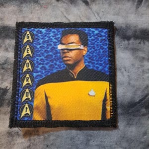 May include: Embroidered patch featuring a portrait of a character from Star Trek: The Next Generation. The patch has a black border and includes the character's name in a vertical column. The character is wearing a yellow uniform and a visor.
