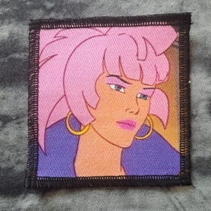 May include: Square patch featuring a cartoon woman with pink hair, gold hoop earrings, and a purple top. The patch has a black border and is set against a gray background. The woman has a serious expression.