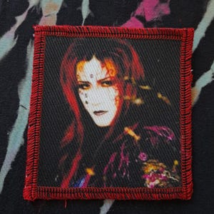 May include: Square patch with a portrait of a person with red hair, pale skin, and dark makeup. The patch has a black background and a red stitched border. The image is a close-up of the person's face and shoulders.