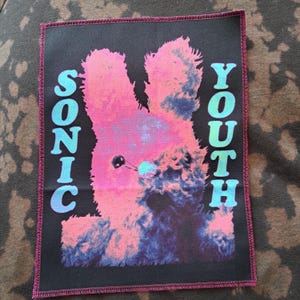 May include: A rectangular patch featuring a pink and blue image of a rabbit-like creature. The words "SONIC YOUTH" are vertically printed in turquoise on either side of the image. The patch has a black background and a pink border.