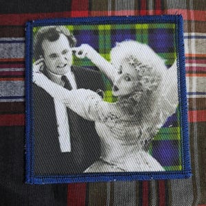 Puede incluir: Parche cuadrado con borde azul. La imagen en blanco y negro muestra a dos personas sobre un fondo de cuadros verdes, azules y amarillos. El parche está sobre una tela a cuadros.
