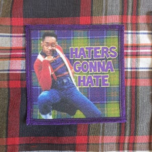 Könnte beinhalten: Quadratischer Aufnäher mit lila Rand, der ein Bild eines Mannes in roter Jacke und blauer Jeans zeigt. Der Text "HATERS GONNA HATE" ist in lila Buchstaben gehalten. Der Aufnäher befindet sich auf einem karierten Stoff.