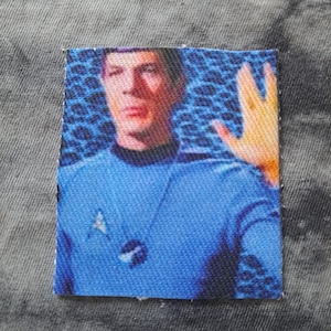 Puede incluir: Parche rectangular con un hombre con uniforme azul, haciendo el saludo vulcano. El parche presenta una insignia de Starfleet. El fondo tiene un patrón azul.