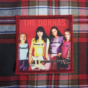 May include: A rectangular patch with a red border and a red background featuring a band photo. The text "THE DONNAS" is at the top. The band members are wearing colourful shirts and holding instruments. The patch is on a plaid fabric.