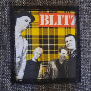 May include: A rectangular fabric patch featuring a yellow and black plaid background with a black and white photograph of four individuals. The word BLITZ is printed in white capital letters on a red rectangular bar at the top of the patch.