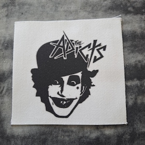 May include: Square, off-white fabric patch featuring a black and white graphic of a smiling face wearing a hat. The text "The Adicts" is above the face. The patch has a slightly textured surface.