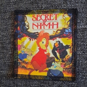 Peut inclure: Un écusson rectangulaire en tissu présentant une illustration colorée de The Secret of NIMH. Le motif montre une souris orange au centre vêtue d'une cape rouge, un corbeau noir et d'autres personnages sur un fond jaune avec du texte noir.