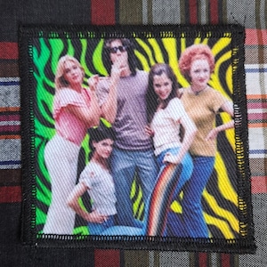 May include: Embroidered patch featuring a group of people in 1970s attire. The patch has a black border and a background with green, yellow, and black zebra stripes. The people are wearing various outfits, including jeans and shirts.