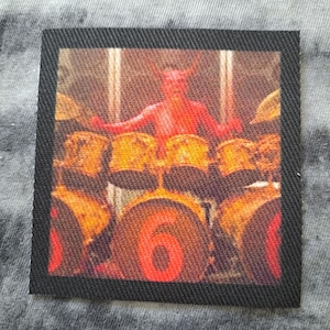 May include: Square patch with a black border featuring a red devil playing a drum set. The number "6" is prominently displayed on the bass drum. The image has a vintage, slightly faded appearance.