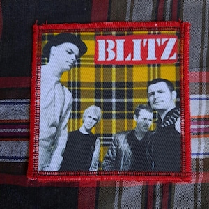 May include: Square patch with a red border featuring a black and white image of a band against a yellow and black plaid background. The word "BLITZ" is in white block letters at the top.