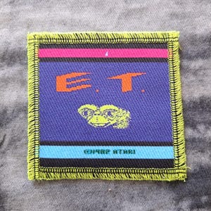 May include: Square patch featuring the letters "E.T." in orange, a green alien, and the text "©1982 ATARI" on a blue background. The patch has a pink and black stripe at the top and a blue stripe at the bottom, with a yellow border.