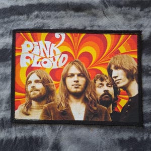 May include: A rectangular patch featuring the band Pink Floyd. The patch has a black border and displays the band members against a vibrant orange and yellow psychedelic background. The band's name is in white text.