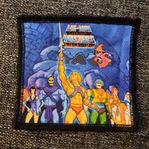 May include: A square fabric patch featuring a colourful graphic of He-Man and the Masters of the Universe characters. The design includes He-Man holding a sword aloft, Skeletor, and other figures against a blue background with the show logo at the top.