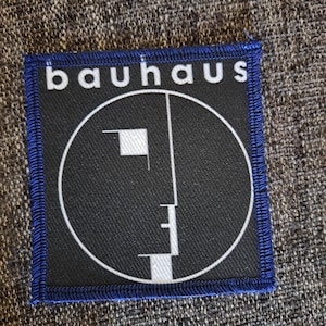 May include: A square fabric patch with a blue stitched border. The centre features a black background with a white circular outline containing a stylised white geometric face profile. The word "bauhaus" is printed in white lowercase letters above.