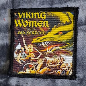 May include: Embroidered patch with the text "Viking Women and the Sea Serpent". The design shows a yellow dragon and figures in a battle scene. The patch has a black border.