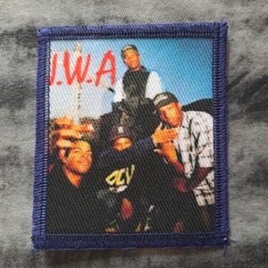 May include: A square patch with a blue border features a group photo with the text "I.W.A." in red and white. The image shows a group of men in various outfits, set against a blue sky background.